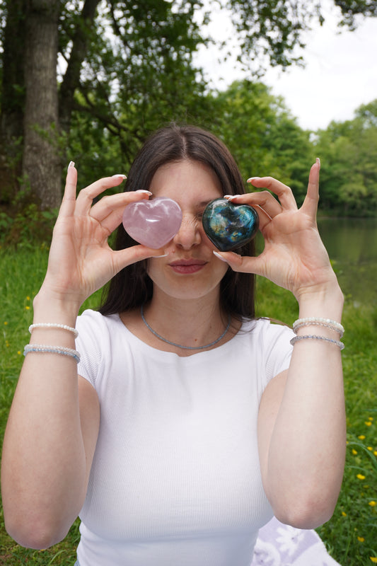 Junge Frau hält ein Rosenquarz- und ein Labradorit-Herz vor ihre Augen, trägt Edelsteinschmuck mit Labradorit, Mondstein und Perlmutt– verspieltes Outdoorshooting mit spirituellem Fokus.