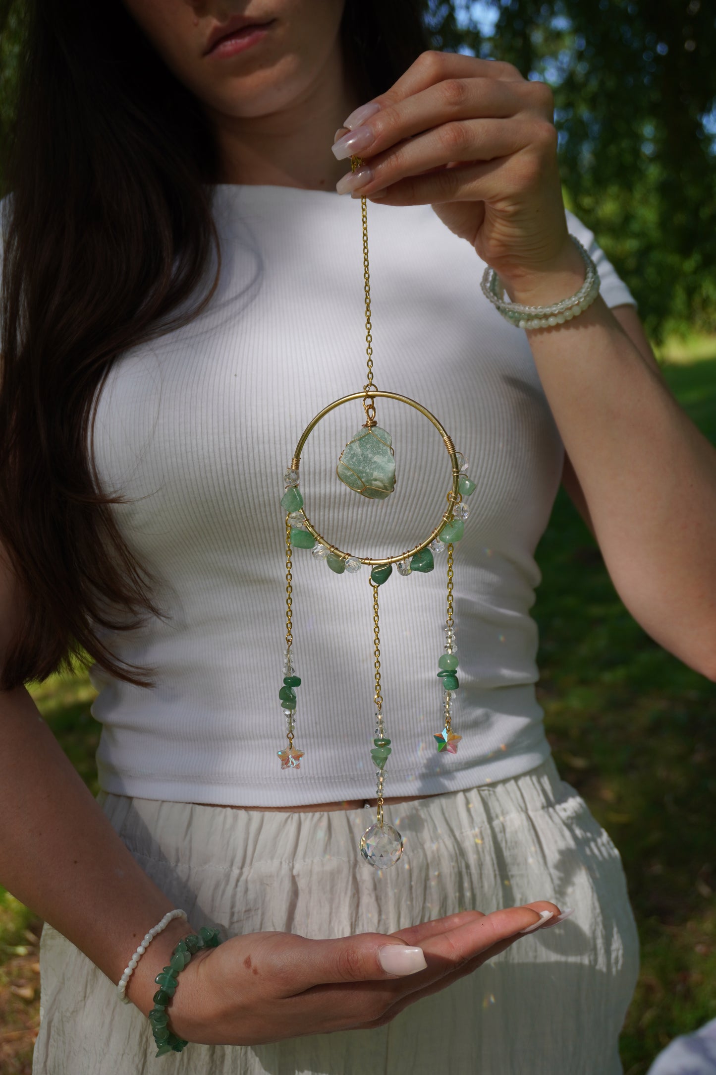 Frau in weißem Outfit präsentiert einen grünen Sonnenfänger mit Aventurin, eingefangen in natürlichem Sonnenlicht unter Bäumen. Sie trägt dabei passenden Edelsteinschmuck. Geschenkidee für besondere Anlässe.