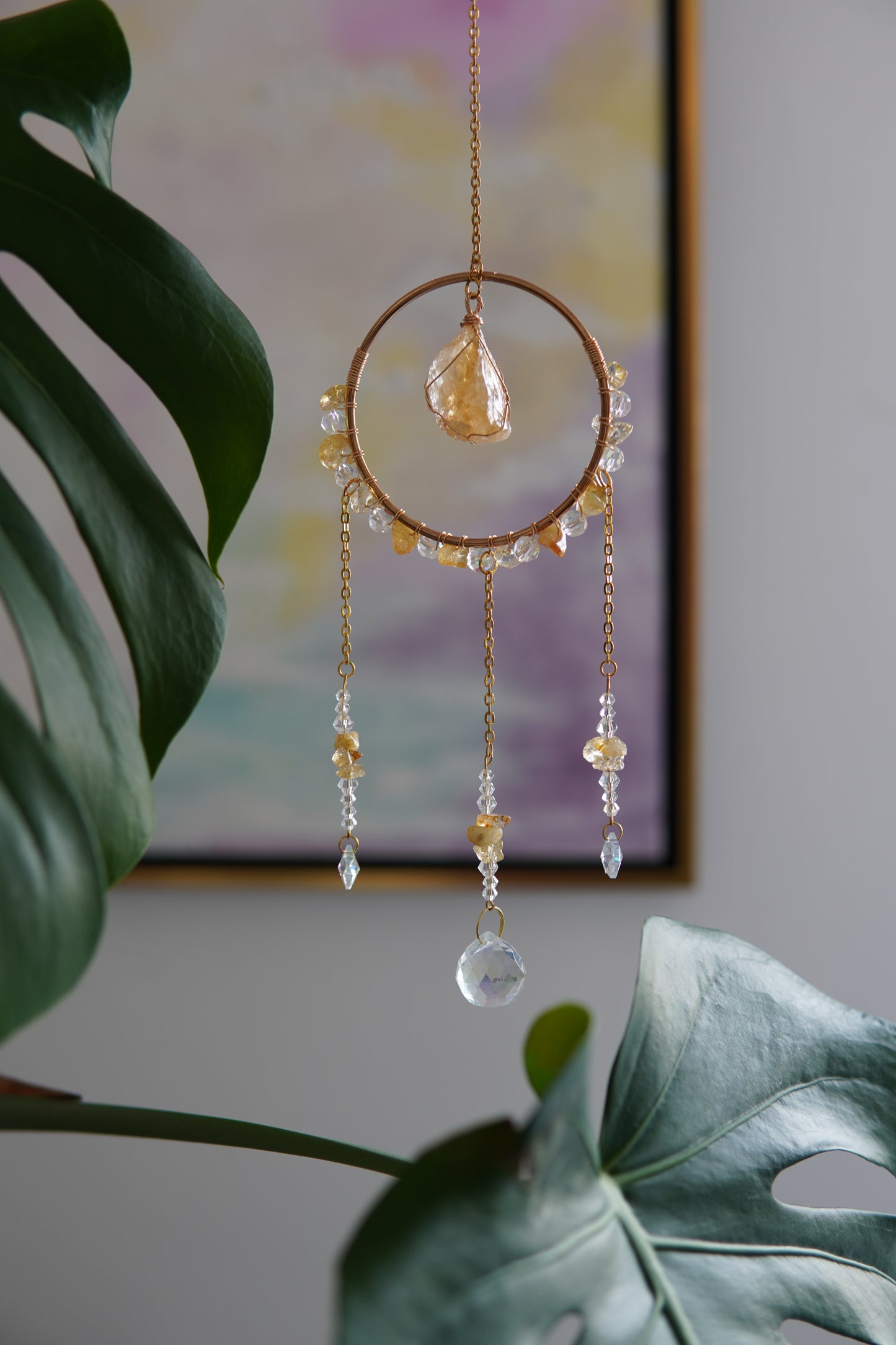 Handgefertigter Sonnenfänger mit goldfarbenem Ring, Citrin-Kristallen und funkelnden Glaselementen, aufgehängt vor einem pastellfarbenen Bild und umgeben von Monstera-Blättern. Wohndekoration. Geschenkidee für besondere Anlässe.