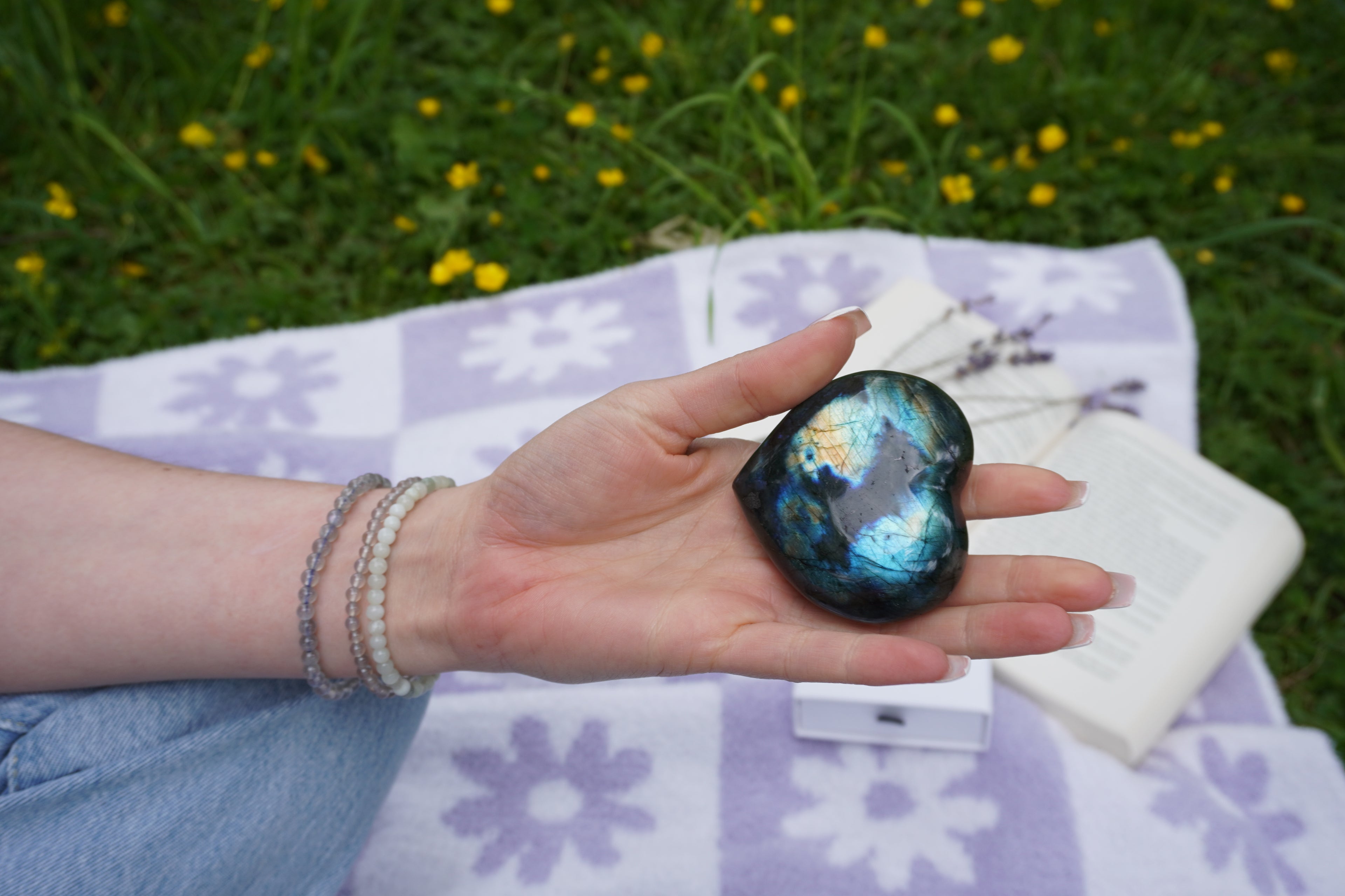 Hand mit Labradorit-Herz auf violett-weißer Picknickdecke, im Hintergrund ein aufgeschlagenes Buch und Lavendel – getragen werden zarte Edelsteinarmbänder aus Jade, Labradorit und Achat.