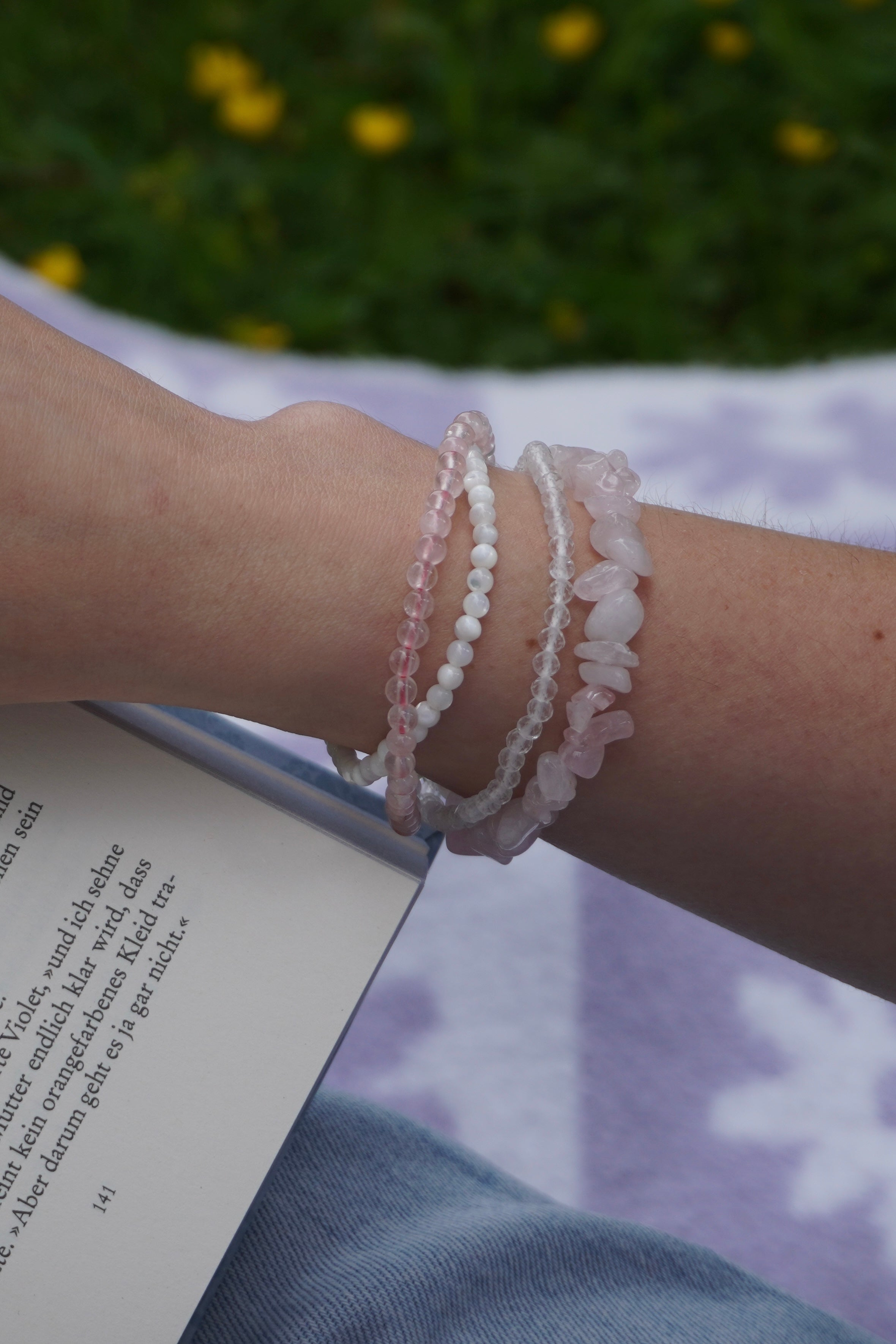 Arm mit vier filigranen Edelsteinarmbändern aus Rosenquarz, Perlmutt und facettiertem Bergkristall – in sommerlicher Lesestimmung auf einer Wiese.