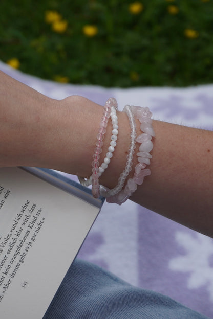 Arm mit vier filigranen Edelsteinarmbändern aus Rosenquarz, Perlmutt und facettiertem Bergkristall – in sommerlicher Lesestimmung auf einer Wiese.