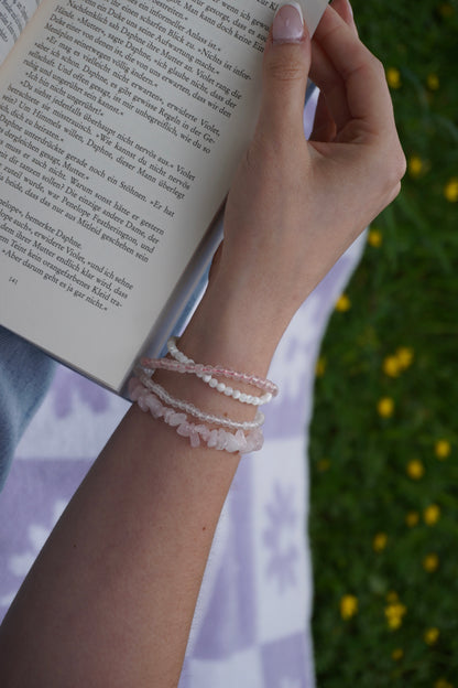 Arm mit vier filigranen Edelsteinarmbändern aus Rosenquarz, Perlmutt und facettiertem Bergkristall – in sommerlicher Lesestimmung auf einer Wiese.