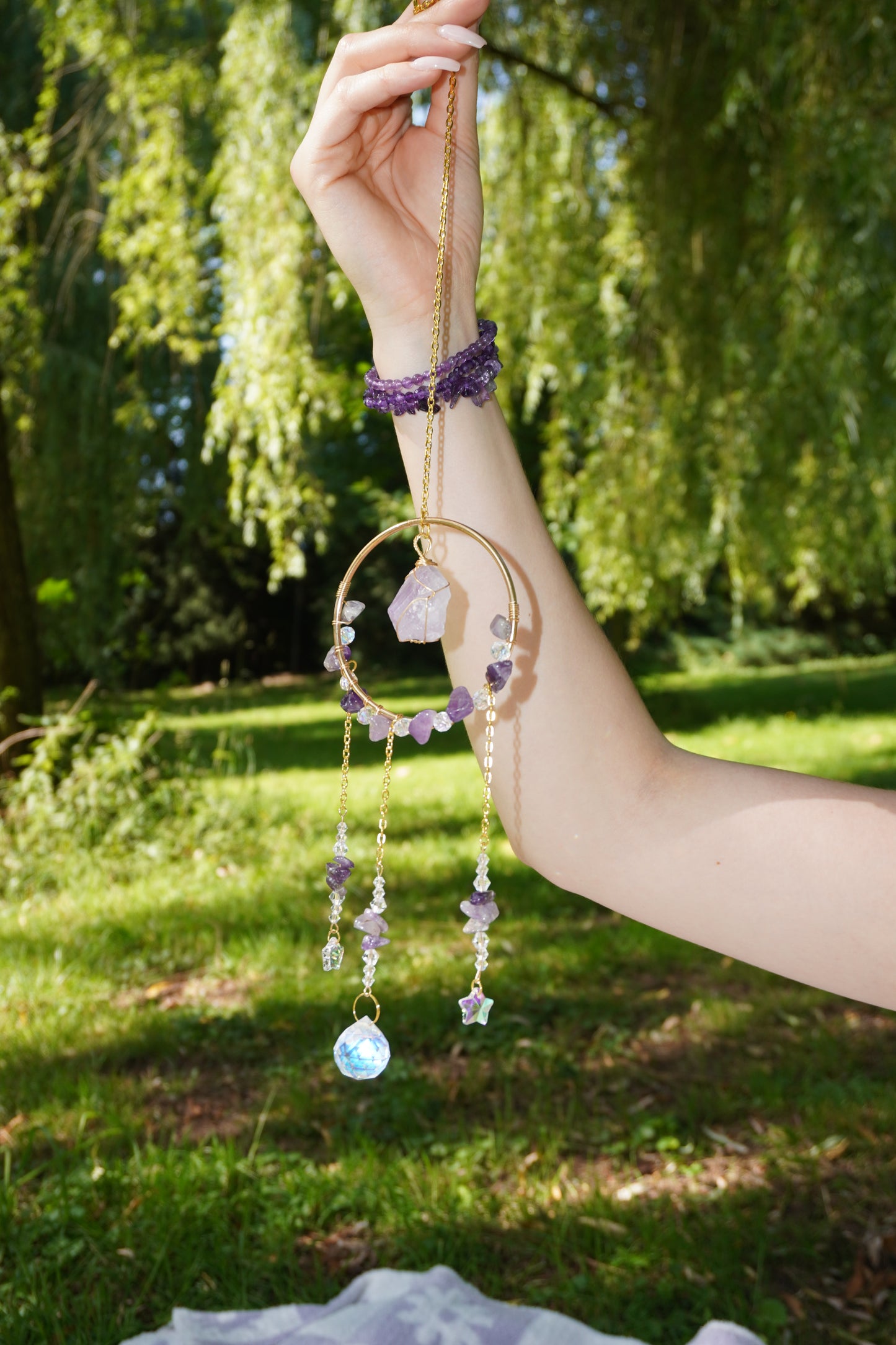 Ein goldener Sonnenfänger mit Amethyst-Splittern und funkelnden Kristallen, gehalten von einer Hand mit passendem Amethystarmband im Grünen.